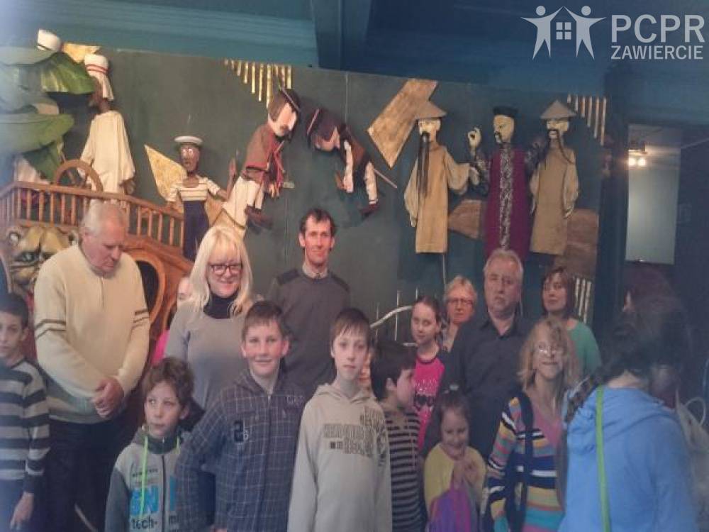 Zdjęcie: Grupa uczestników i uczestniczek projektu wraz z dziećmi stojąca we wnętrzu Teatru, a w tle kukiełki z przedstawień